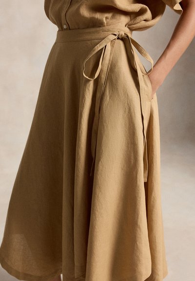 Moteris dėvi smėlio spalvos įvyniojamą sijoną, surištą ties juosmeniu, dešine ranka kišenėje ant neutralios foninės sienos.