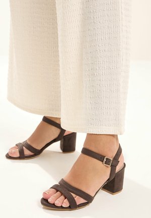 Fødder iført mørkebrune sandaler med blokhæl og ankelsremme, kombineret med teksturerede hvide bukser med vide ben.