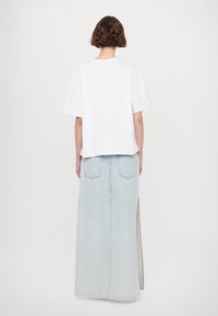 Personne aux cheveux bruns courts portant un t-shirt blanc uni à manches courtes et une jupe longue en denim bleu clair avec une fente latérale, vue de dos.