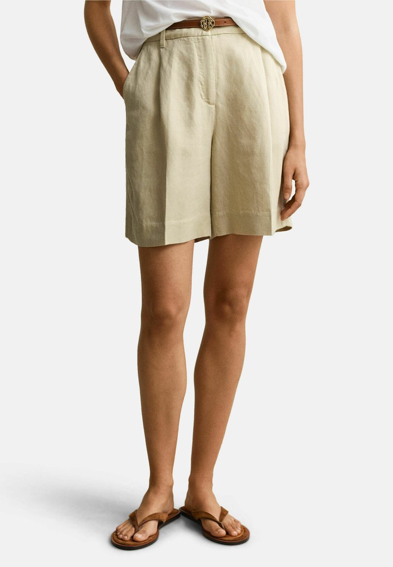 Personne portant un short beige mi-long avec des poches, un haut blanc rentré, une ceinture marron avec une boucle circulaire, et des sandales marron.