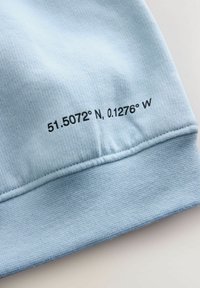 Hele sinine kangas, sileda tekstuuriga, millel on musta värvi trükitud koordinaadid: "51.5072° N, 0.1276° W" ääre lähedal.