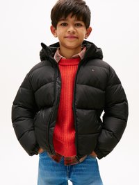 Veste matelassée noire avec une capuche, dotée d'une fermeture éclair et d'un logo. Portée sur un pull en maille rouge et une chemise à carreaux, associée à un jean bleu.