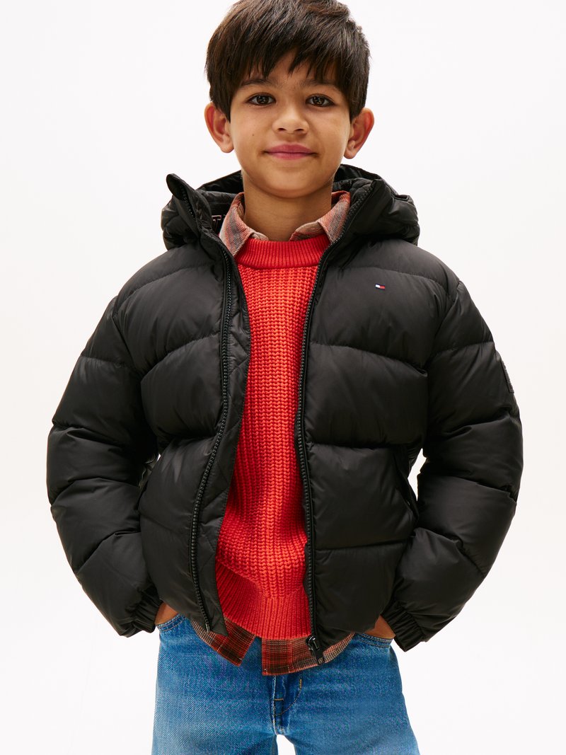 Veste matelassée noire avec une capuche, dotée d'une fermeture éclair et d'un logo. Portée sur un pull en maille rouge et une chemise à carreaux, associée à un jean bleu.