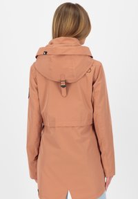 Giacca impermeabile marrone chiaro con cappuccio, ventilazione posteriore, vita regolabile e tasche laterali. Presenta un branding minimale e una texture liscia.