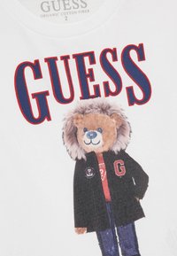 T-shirt bianco con un personaggio orso dalla pelliccia beige, che indossa una giacca nera, una maglietta rossa e jeans blu, con il testo "GUESS" sopra.