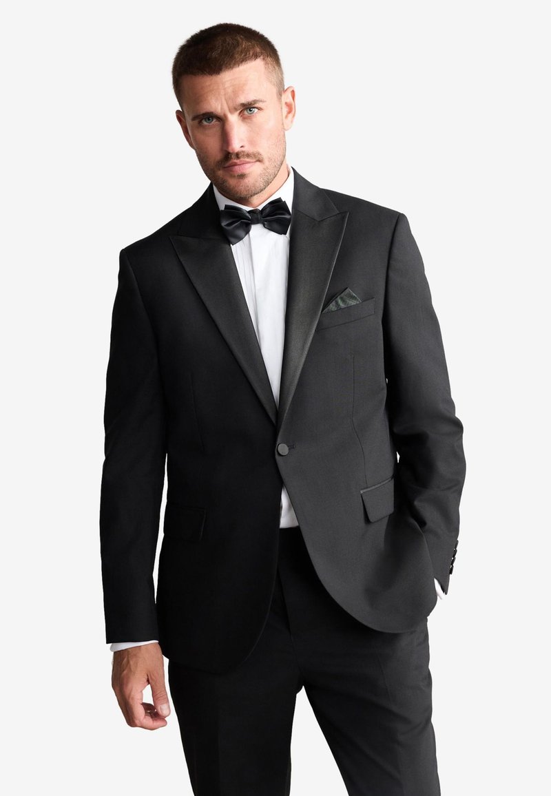 Homme aux cheveux courts et à la barbe taillée, portant un smoking noir à revers en satin, une chemise blanche et un nœud papillon noir, debout avec une main dans la poche.