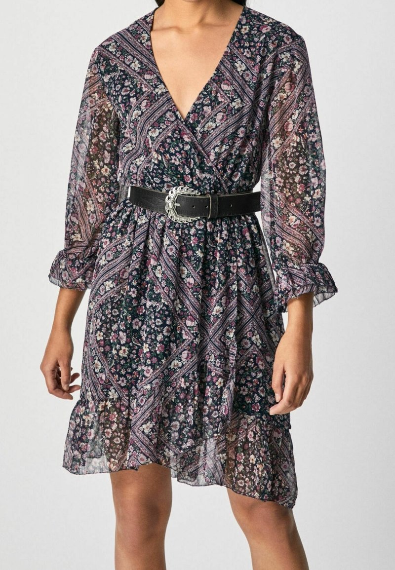 Femme portant une robe portefeuille noire à motifs floraux roses et blancs, manches longues transparentes, ourlet volanté et ceinture noire avec boucle argentée.