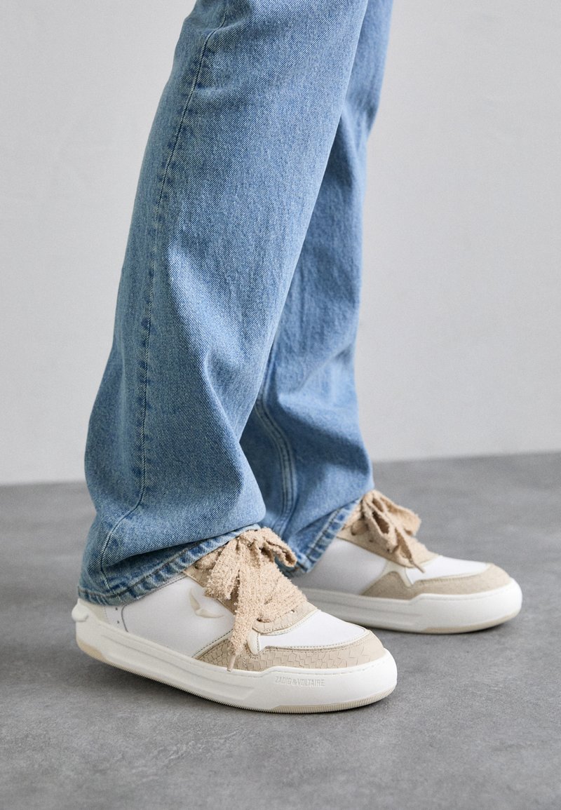 White and beige sneakers with textured panels, featuring beige laces and a rubber sole, paired with light blue denim jeans.