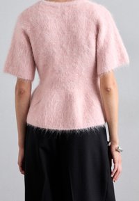 Růžový huňatý svetr s krátkými rukávy, zúženým pasem a měkkou strukturou. V kombinaci s černou sukní, která ukazuje kontrastní barvy a materiály.