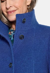 Blauer Wollmantel mit hohem Kragen, der über drei große schwarze Knöpfe, eine strukturierte Verarbeitung und subtile Nähdetails verfügt.