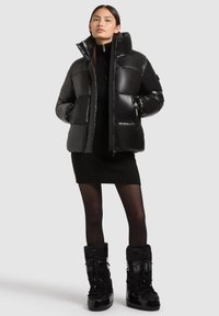 Melnā pūkaini jaka ar augstu apkakli, mirdzošu tekstūru un ar rāvējslēdzēju priekšā, apvienota ar ribotu melnu kleitu un melniem sniega zābakiem.