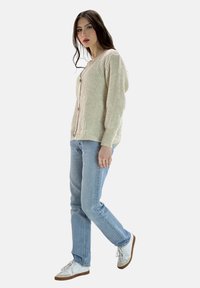 Beige gerippte Strickjacke mit Knopfverschluss, getragen über hellblauen Jeans mit geradem Bein. Das Modell steht vor einem neutralen Hintergrund und präsentiert die Details des Outfits.