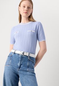 Lys blå strikket topp med korte ermer, med hvit kant og to brystlommer med knapper. Kombinert med høyt liv, blå jeanstwill og et hvitt belte.