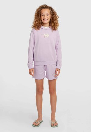 Naeratav lokkis juustega tüdruk kannab lavendlivärvi kapuutsi ja sobivaid lühikesi pükse logodega, seistes paljajalu heledal taustal.