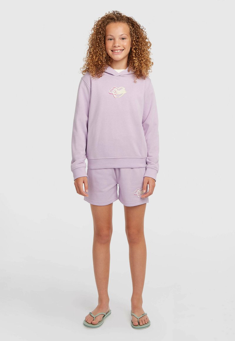Fille souriante aux cheveux bouclés portant un sweat à capuche lavande et un short assorti avec des logos, debout pieds nus sur un fond clair uni.