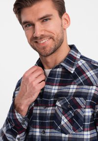 Camicia in flanella a quadri blu navy e rossa con colletto, due tasche sul petto e chiusura a bottoni. Texture morbida con un design dalla vestibilità rilassata.