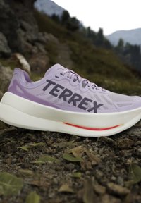 Μωβ παπούτσι για τρέξιμο σε μονοπάτι Terrex με παχιά λευκή σόλα σε βραχώδες ορεινό περιβάλλον με θολό φόντο.