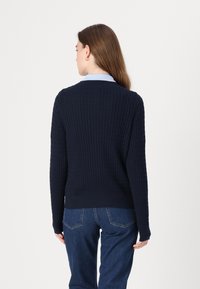 Σκούρο μπλε πλεκτό πουλόβερ με υφή, στρογγυλή λαιμόκοψη και ριμπ τελείωμα. Φοράει πάνω από ένα ανοιχτό μπλε πουκάμισο με γιακά, με μακριά μανίκια.