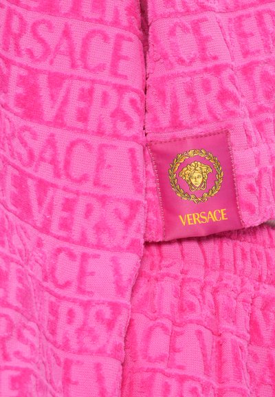 Rožna kopalna plašča z reliefnim vzorcem logotipa Versace. Ima mehko teksturo in izrazito blagovno oznako z zlatimi detajli.