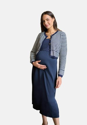 Tehotná žena v námorníckych modrých šatách a so vzorovaným svetrom jemne drží svoj bruško a usmieva sa na jednoliatom pozadí.