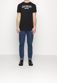 Sort bomulds t-shirt med "GUESS EST. 1981" trykt i fede hvide og lilla bogstaver, parret med blå jeans og sorte læderstøvler.