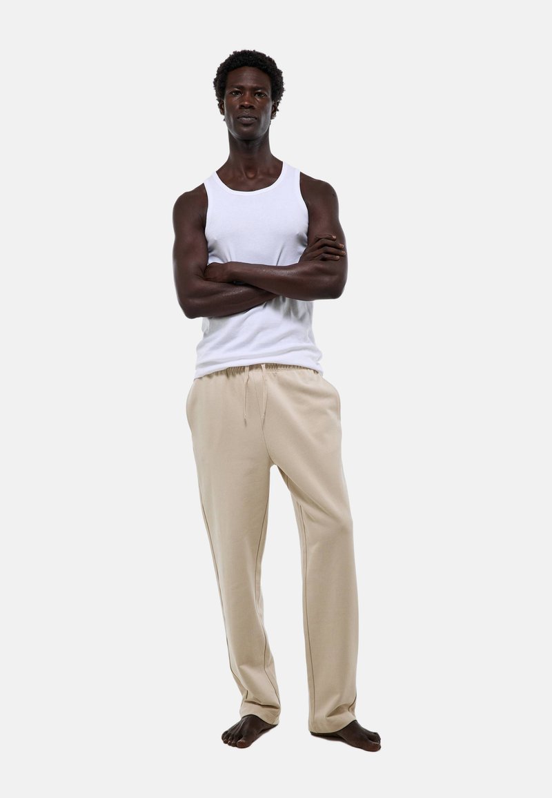 Un débardeur blanc et un pantalon de jogging beige, avec une taille à cordon de serrage, un tissu doux et une coupe décontractée. Le modèle se tient pieds nus, les bras croisés.