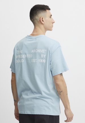 Light blue cotton t-shirt featuring white text on the back: “THE ARCHIVE,” “PRESENTED BY,” “SOLID EST. 1999.” Short sleeves, relaxed fit.