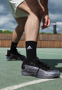 Sorte treningssko med en teksturert sølvfarge panel på siden og oransje detaljer, kombinert med sorte sokker med Adidas-logo, på en basketballbane.