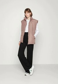 Jovem mulher de pé, vestindo um colete matelassado mauve, uma blusa branca de mangas longas, calças largas pretas e ténis brancos com detalhes verdes.