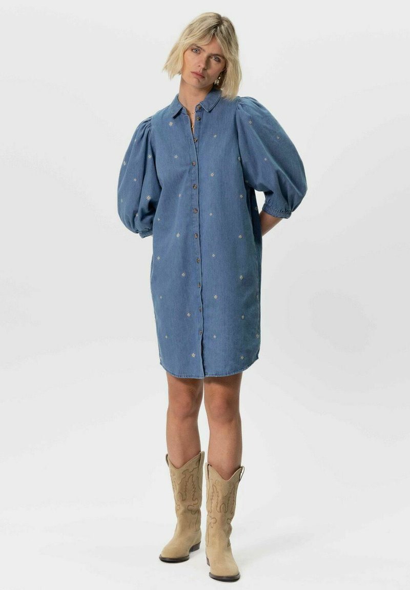 Robe chemise en denim avec des manches bouffantes courtes, des boutons sur le devant et une broderie florale. Portée avec des bottes de cowgirl beiges, longueur genou, coupe décontractée.