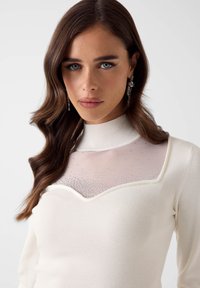 Witte trui met een hoge halslijn, voorzien van een transparante mesh inzet met kleine, verspreide strassteentjes als accent.