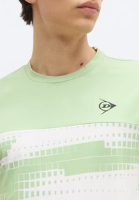 Άνδρας που φοράει ανοιχτό πράσινο και λευκό πουκάμισο με σχέδιο πλέγματος και μικρό μαύρο λογότυπο στο στήθος, κοιτάζοντας ελαφρώς προς τα πλάγια.