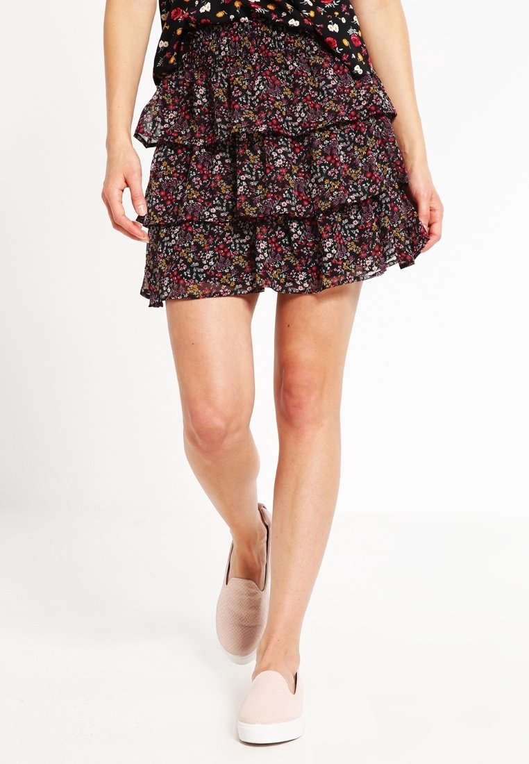 Falda negra con flores, de capas acanaladas y volantes, con pequeños dibujos de flores en rojo, amarillo y blanco. Combinada con unos zapatos de lona rosas claros.