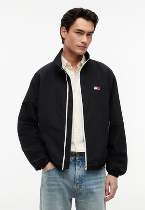 Junger Mann mit dunklen Haaren, der eine schwarze Jacke mit Reißverschluss über einem hellen Hemd und ausgewaschenen blauen Jeans trägt, steht mit den Händen in den Taschen.