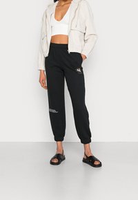 Lätt beige zip-jacka över en vit crop-top, tillsammans med svarta joggers med logodetaljer och svarta slip-on sandaler.