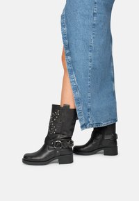 Bottines en cuir noir avec des clous décoratifs, des accents de boucle en métal et un talon épais, associées à une jupe en denim avec des fentes sur les côtés.