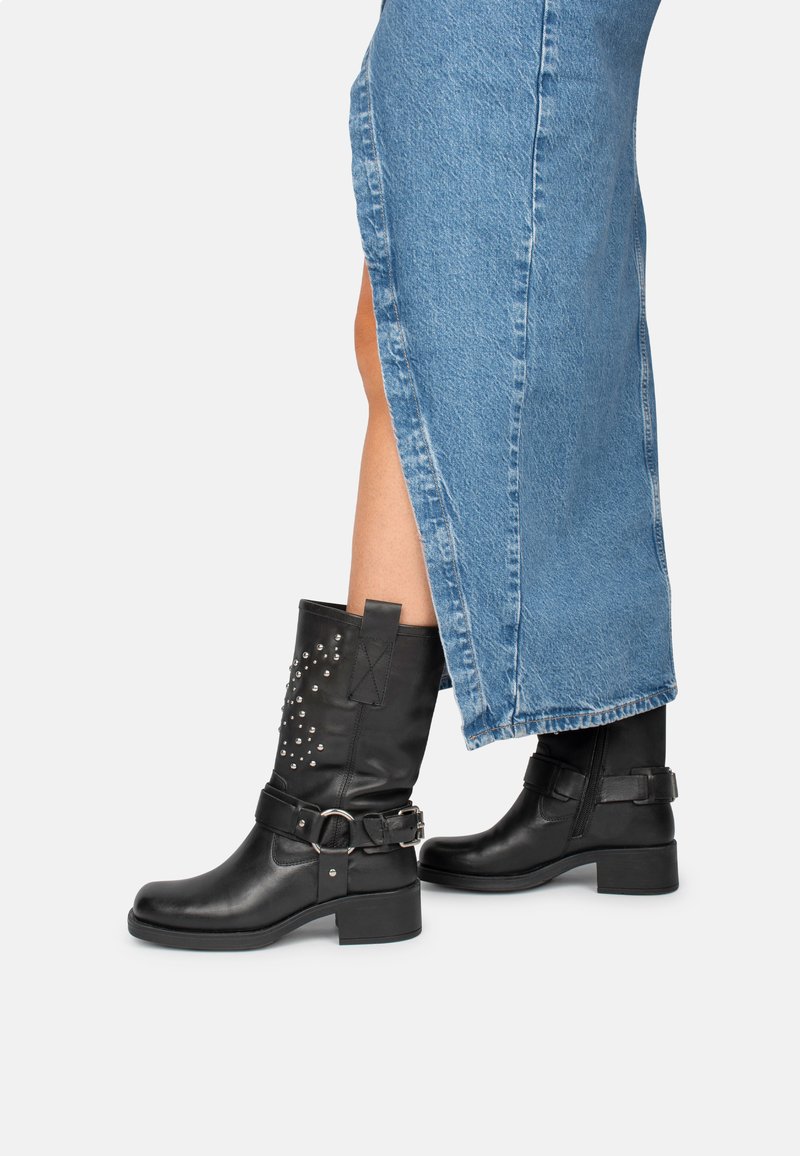 Bottines en cuir noir avec des clous décoratifs, des accents de boucle en métal et un talon épais, associées à une jupe en denim avec des fentes sur les côtés.