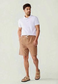 Shorts marron à côtes avec une taille à cordon, associés à un polo blanc. Les chaussures sont des sandales beige avec des sangles réglables.