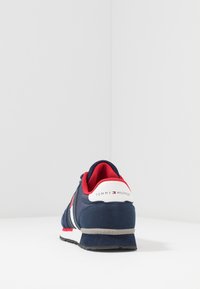 Mörkblå sneaker med röda och vita detaljer, försedd med en ovandel i mocka, vadderad krage och texturerad gummisula. Logotyp på hälfliken.