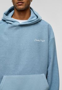 Lichtblauwe hoodie van zacht, textuurstof, met een voorvak en "Eternity Project" geborduurd in het wit op de borst.