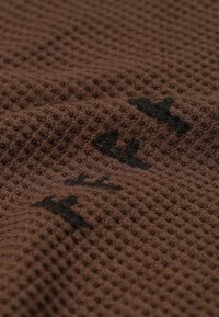 Tissu texturé brun avec un motif en gaufre, présentant des accents imprimés noirs. Le matériau semble doux et flexible.