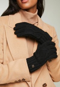 Mustmustest mustriibulised kindad, millel on kaabelmustristik ja soonikääred, millel on kuldne detail, koos beežist mantli ja kõrge kaelusega särgiga.
