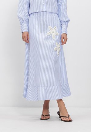 Vrouw draagt een midi-rok met blauw-witte strepen en twee witte bloemenapplicaties, gecombineerd met een bijpassend gestreept overhemd en zwarte sandalen met hak.