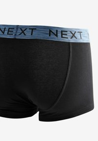 Melnas kokvilnas bokseršortus ar zilu svītrotu jostu, kurā ir teksts "NEXT" melnā krāsā. Gluda tekstūra ar minimālu šuvju skaitu.