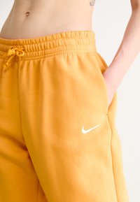 Plano cercano de la cintura de una persona que lleva pantalones de chándal amarillos mostaza de Nike con cinturilla elástica y una mano en el bolsillo.