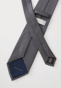 Cravate grise à motif avec un tissu texturé, doublée de marine et étiquetée "Michael Kors". Motif géométrique sur toute la cravate.