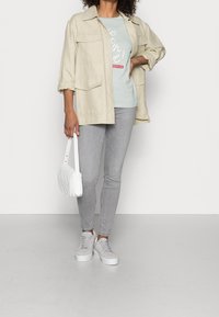 Chaqueta oversized beige clara con múltiples bolsillos, camiseta gráfica verde menta, vaqueros slim grises, bolso texturizado blanco y zapatillas grises.