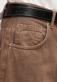 Τσέπη καφέ τζιν με μεταλλικά καρφιά και μαύρη δερμάτινη ζώνη περασμένη από τις θηλιές της ζώνης.