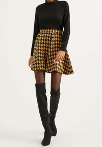 Mujer vistiendo un top negro de manga larga, una falda de cuadros marrón y negro, medias negras transparentes y botas altas negras contra un fondo blanco.