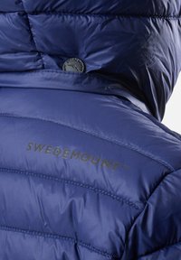 Navy blue puffy jacket with horizontal quilted design, featuring a black logo reading "SWEDEMOUNT" near the neckline and smooth texture.